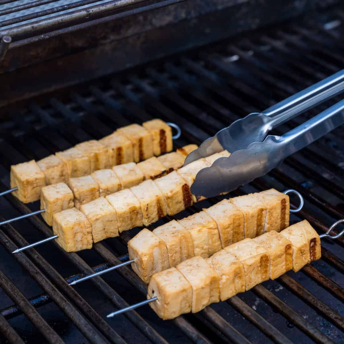 Tongs flipping tofu skewers on an outdoor gas grill.