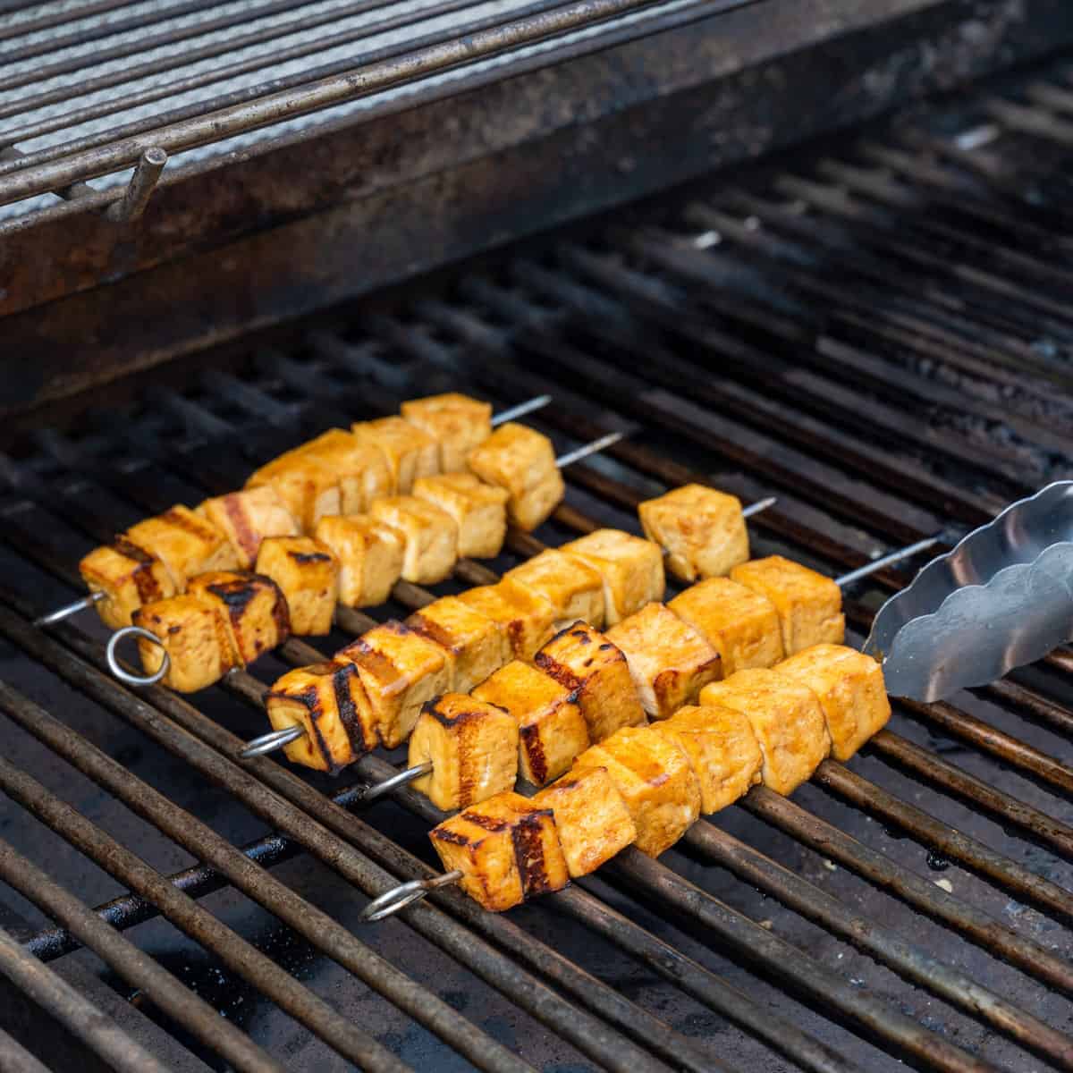 Tongs flipping marinated grilled tofu skewers on an outdoor gas grill.