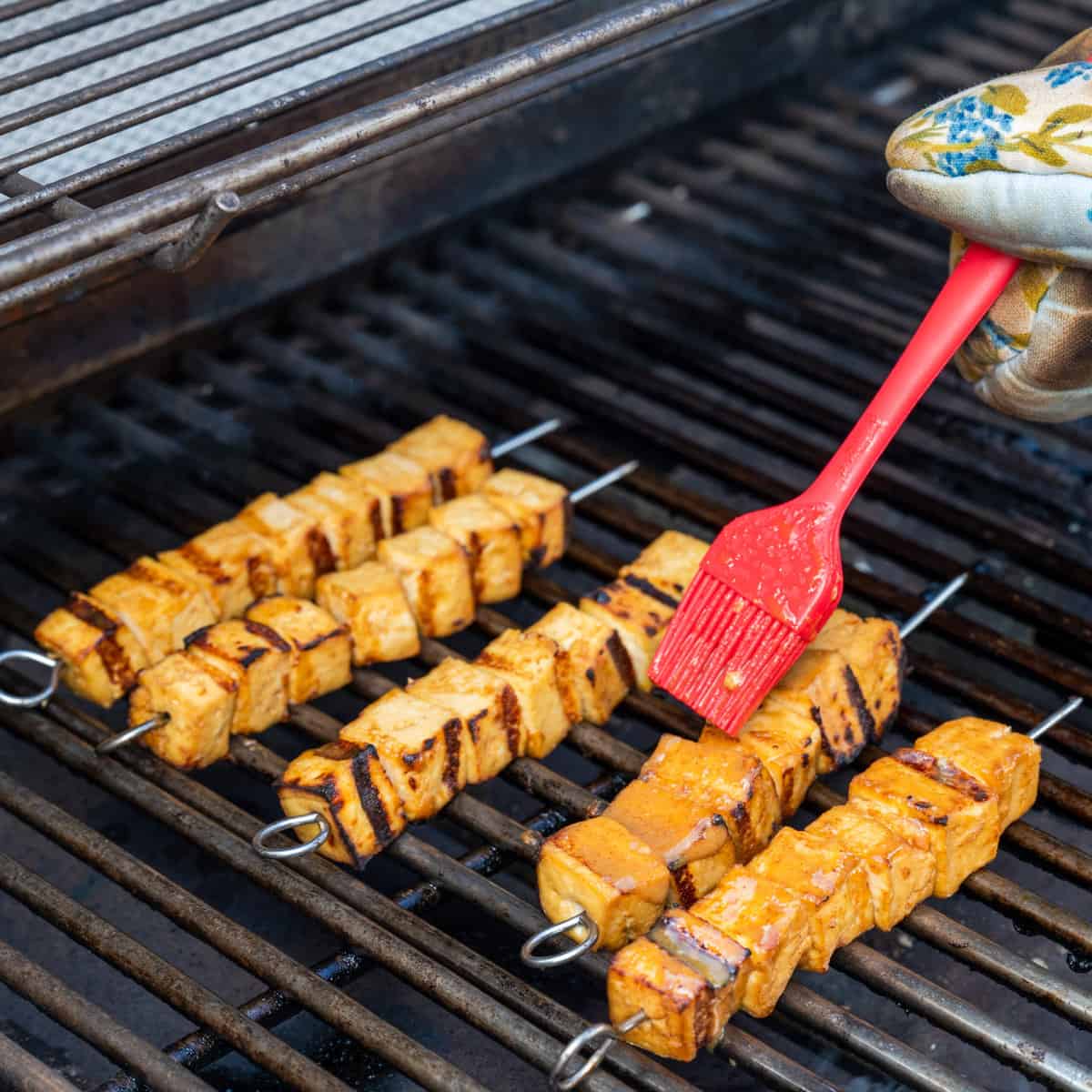 Brushing marinade onto grilled tofu skewers on an outdoor gas grill.