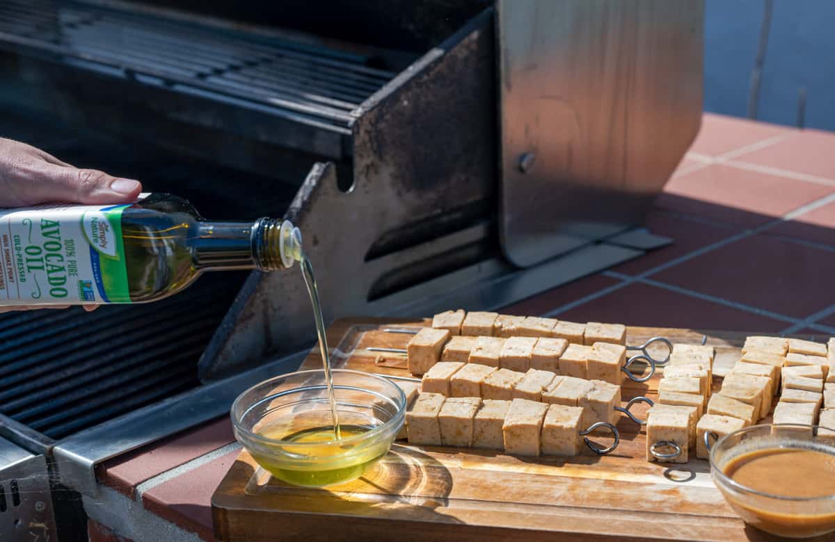 pouring avocado oil into a bowl next to tofu skewers about to be grilled outside.