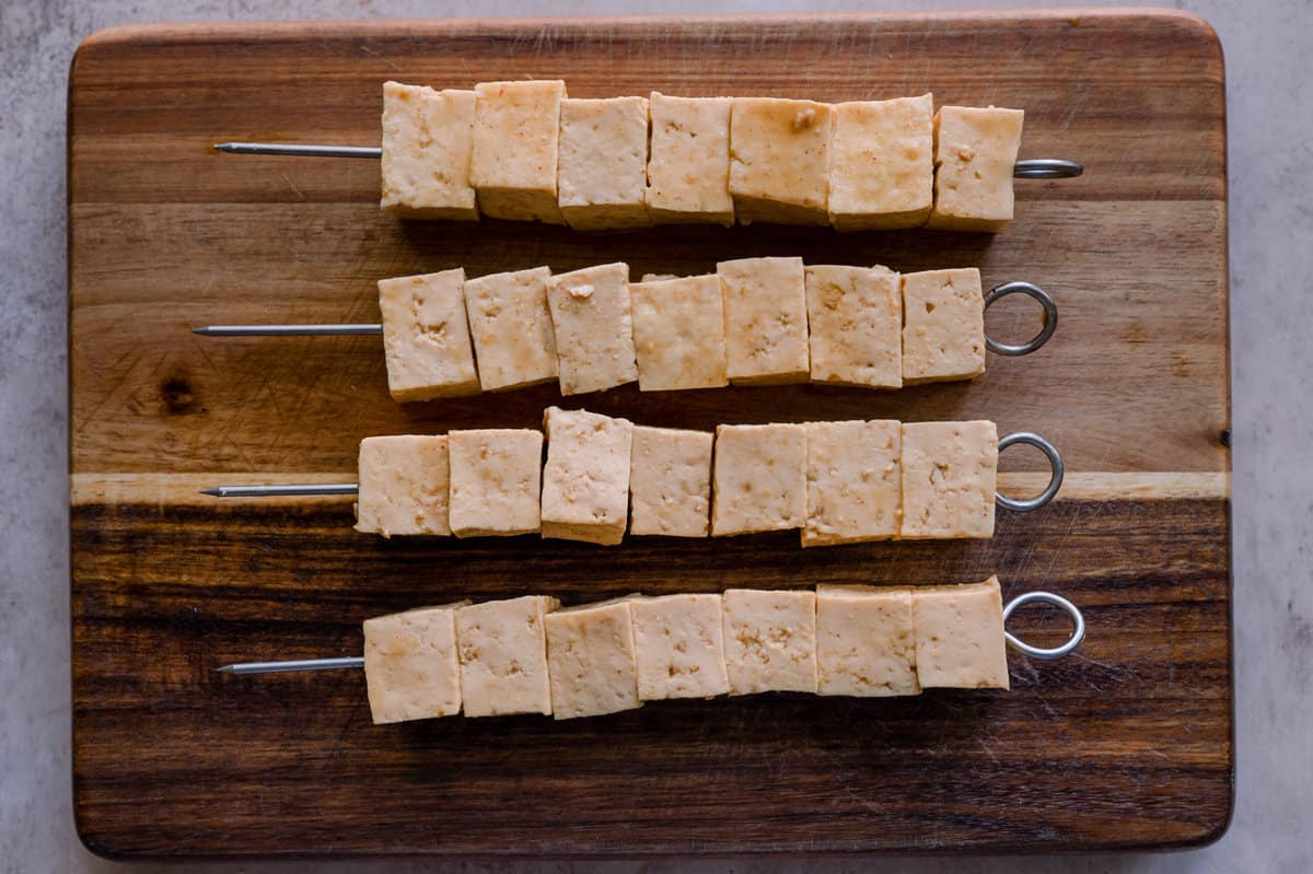 marinated tofu cubes threaded on metal skewers, sitting on a wooden cutting board.