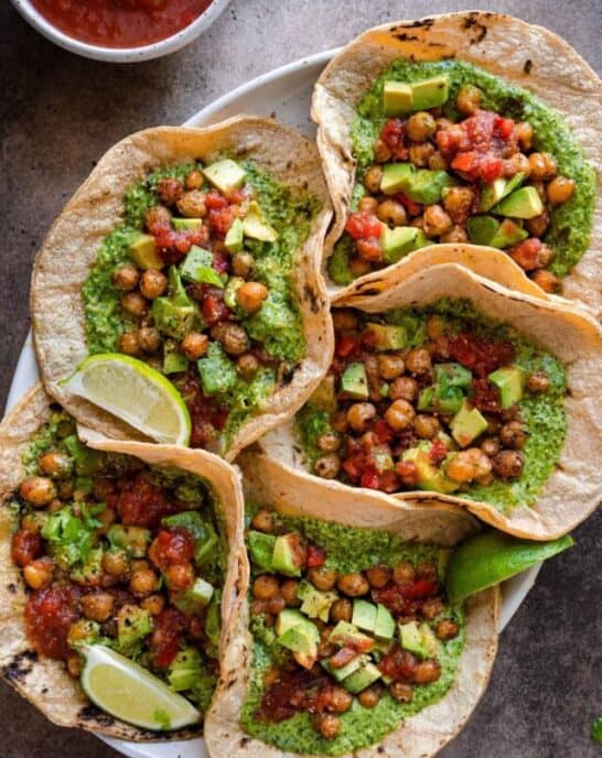 vegan chickpea tacos with cilantro pesto served with salsa and avocado, five tacos arranged on a platter on a brown backdrop.