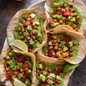 vegan chickpea tacos with cilantro pesto served with salsa and avocado, five tacos arranged on a platter on a brown backdrop.
