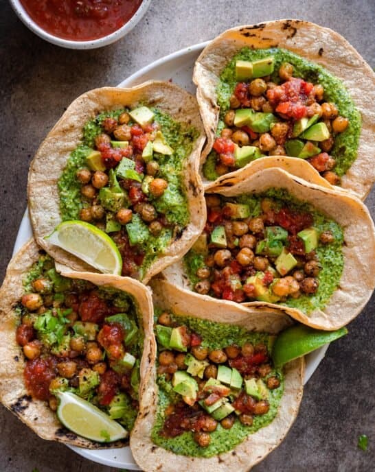 vegan chickpea tacos with cilantro pesto served with salsa and avocado, five tacos arranged on a platter on a brown backdrop