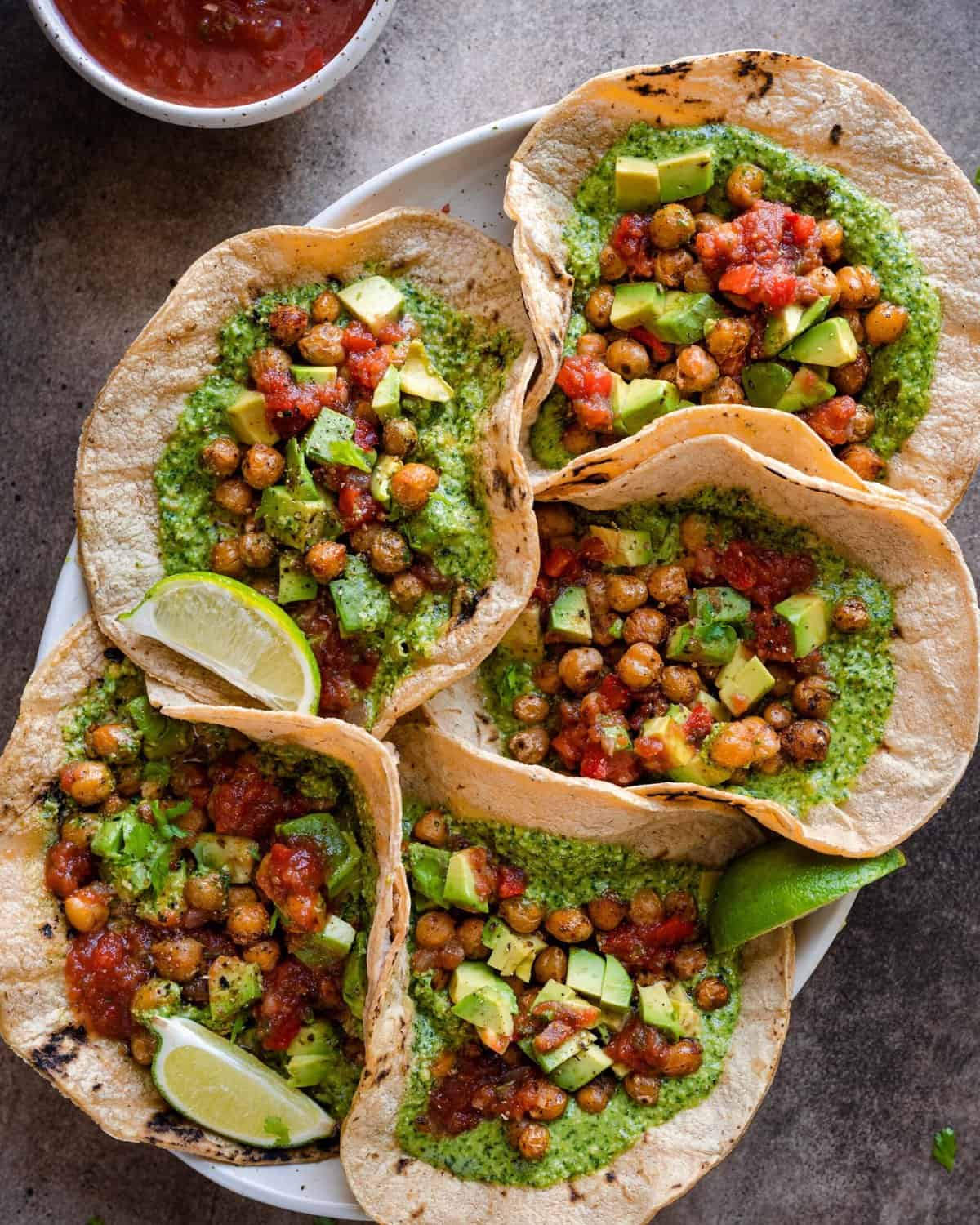 vegan chickpea tacos with cilantro pesto served with salsa and avocado, five tacos arranged on a platter on a brown backdrop