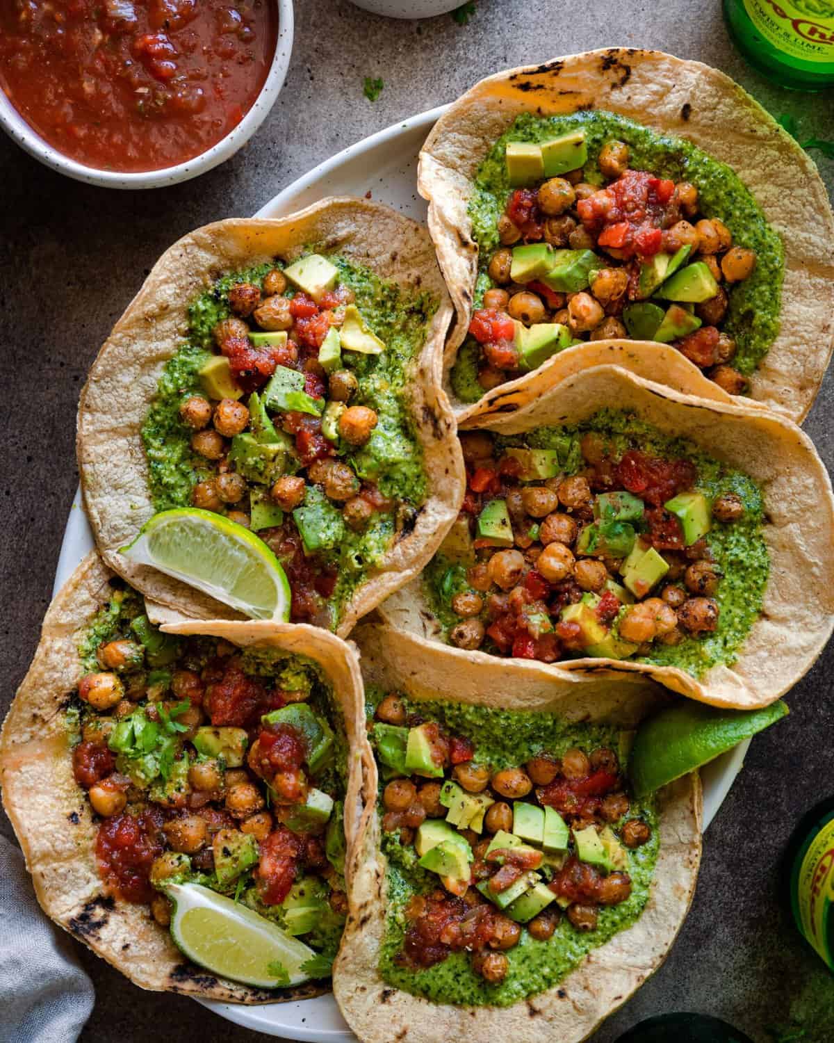 vegan chickpea tacos with cilantro pesto served with salsa and avocado, five tacos arranged on a platter on a brown backdrop