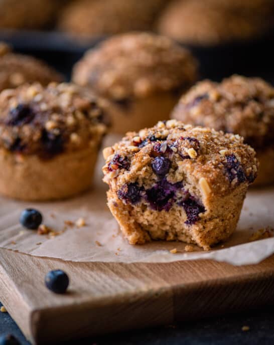 vegan blueberry muffin with bite shot on parchment paper with other muffins in background.