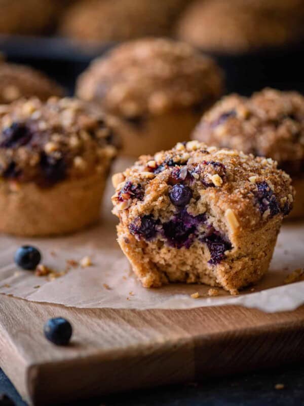 vegan blueberry muffin with bite shot on parchment paper with other muffins in background.