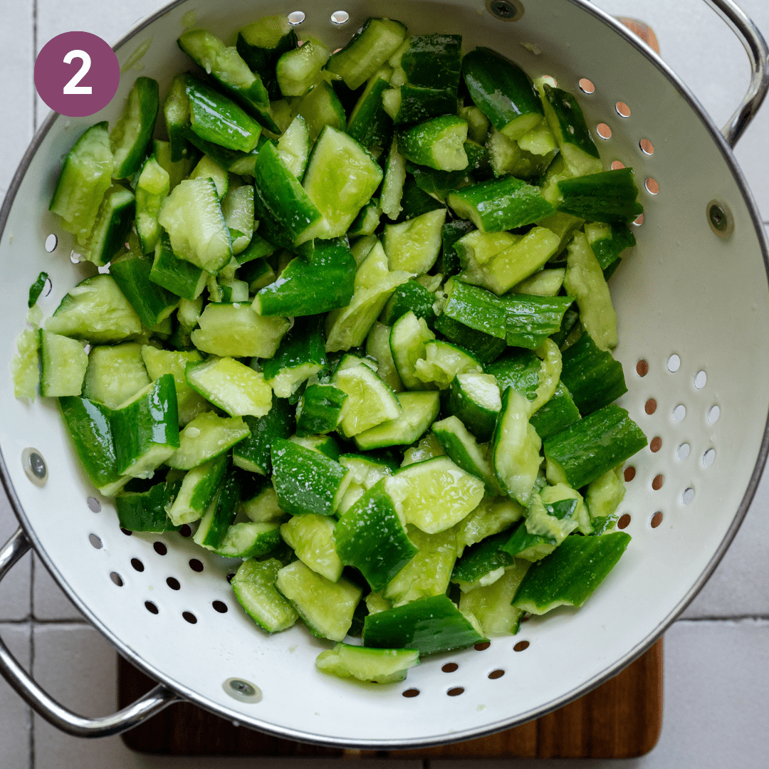 smashed cucumbers with salt in a white colander on a wooden cutting board.