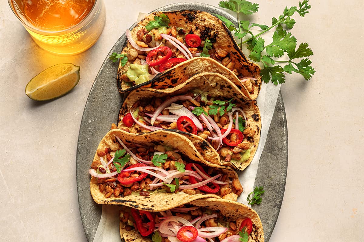 Platter of four tempeh tacos with pickled shallots, next to a glass of beer on a table.