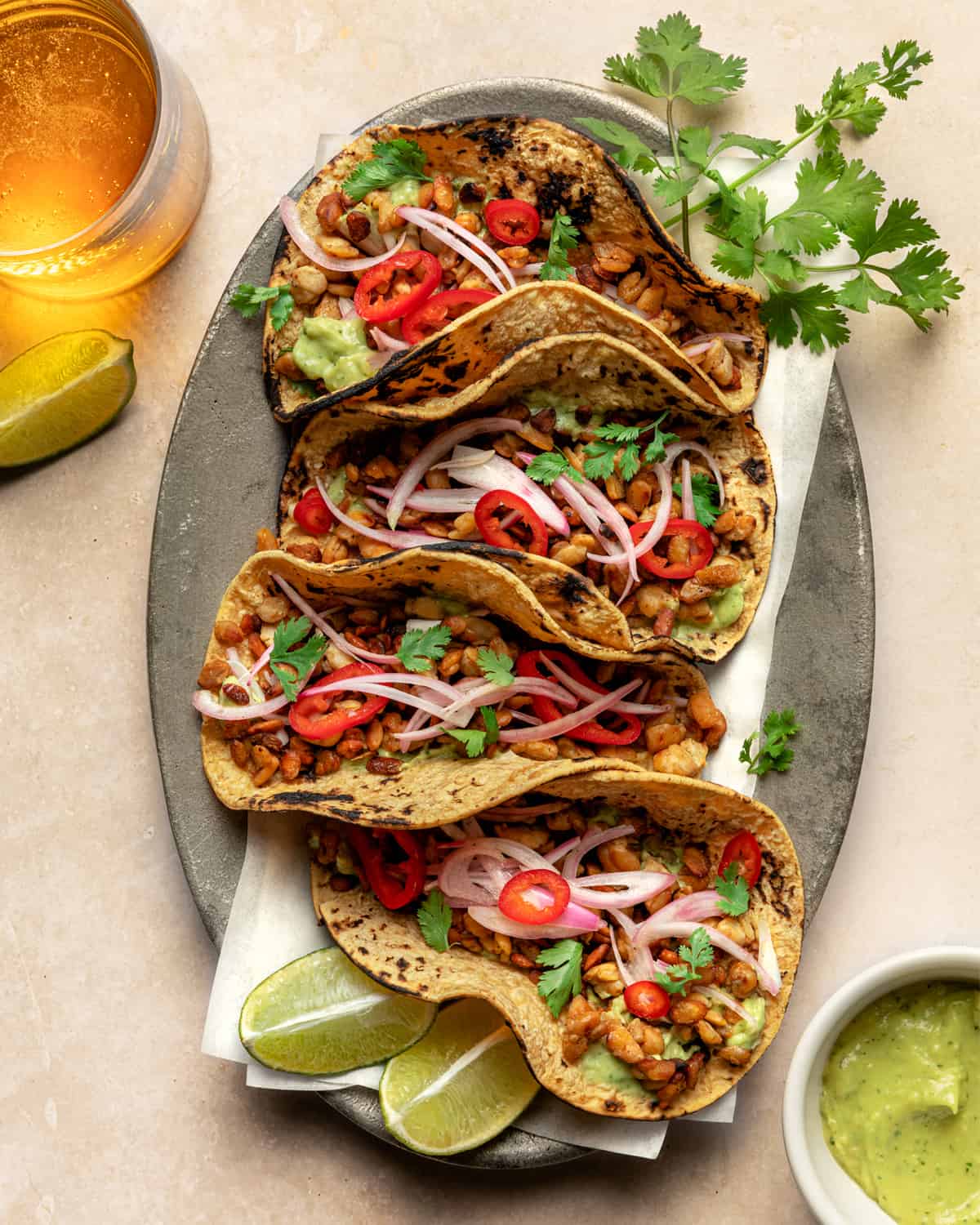 A large platter with four tempeh tacos, garnished with cilantro with glass of beer. 