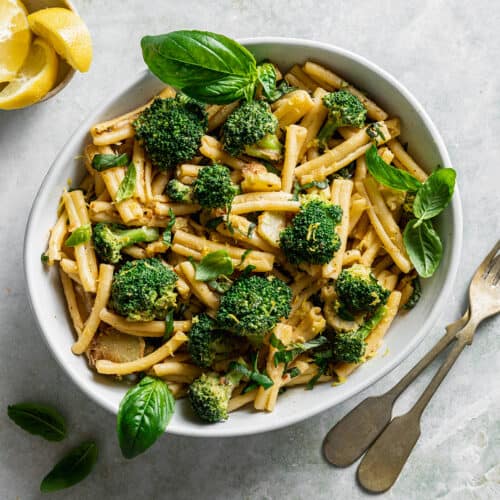 a ceramic bowl filled with tahini pasta and broccoli topped with basil on a marbled surface.