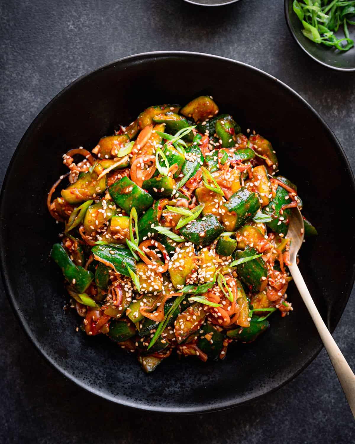 spicy korean cucumber salad in a black bowl with a gold spoon, on a dark grey surface.