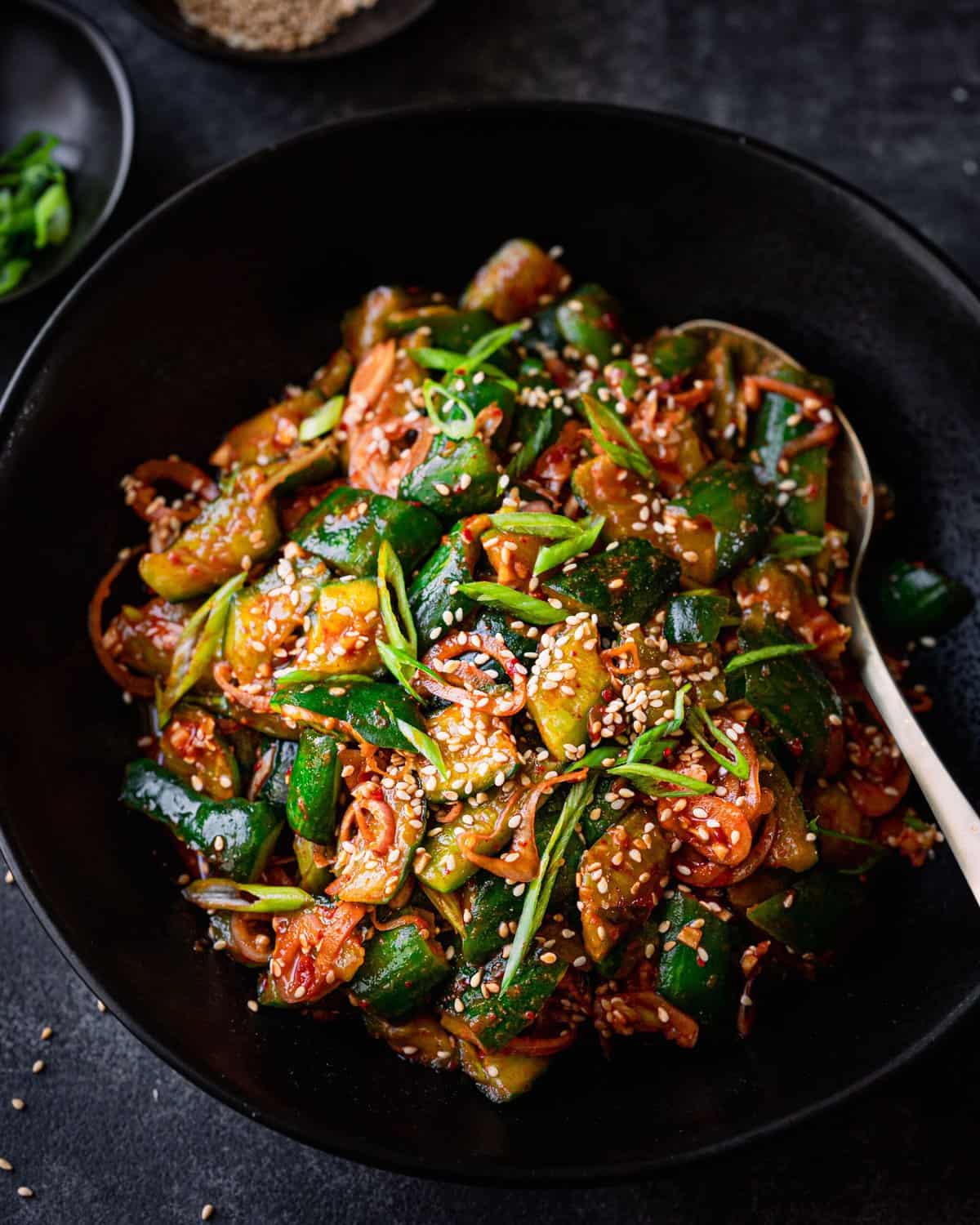 spicy korean cucumber salad in a black bowl with a gold spoon, on a dark grey surface.