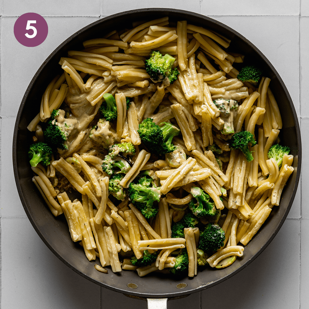 tahini pasta mixed with broccoli in a frying pan on a white titled surface. 
