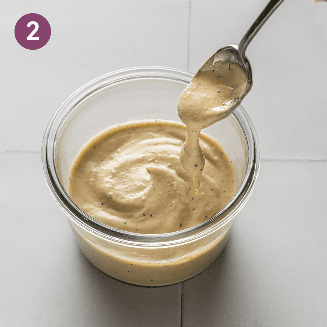 spoon stirring tahini sauce in a glass jar on a white tiled surface. 