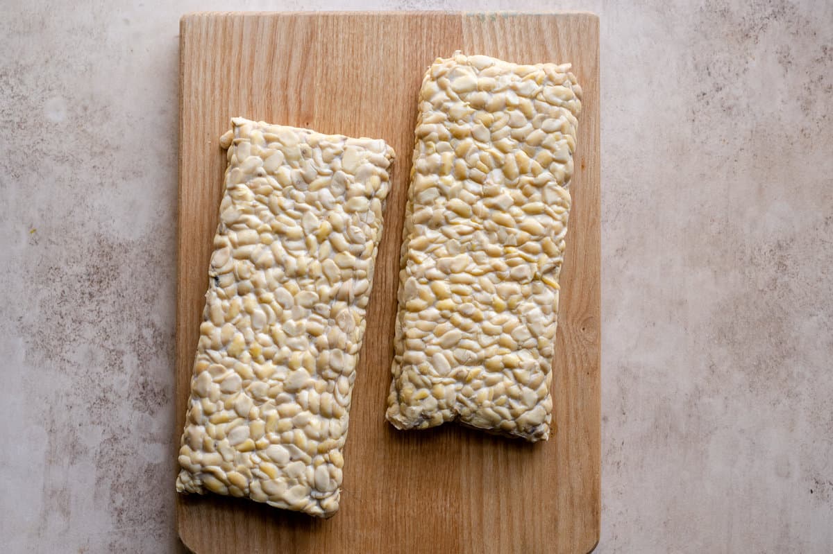 two blocks of tempeh on a wooden cutting board on a pink surface.