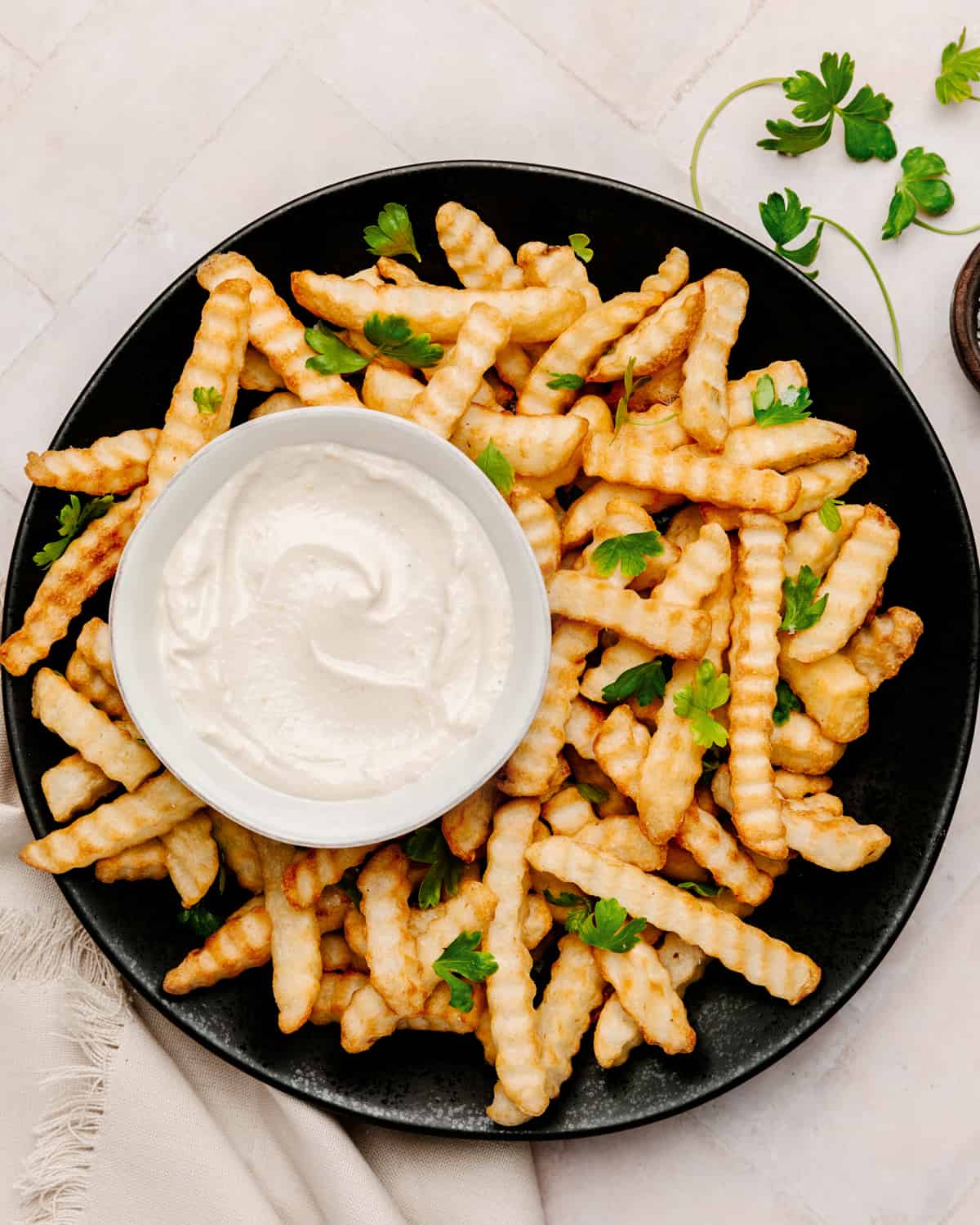 Large black plate with crinkle-cut fries topped with parsley and a bowl of vegan aioli.