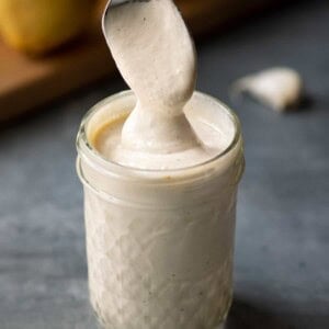spoon dipping into jar of vegan aioli, with lemon and garlic in the background.