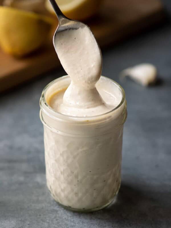 spoon dipping into jar of vegan aioli, with lemon and garlic in the background.