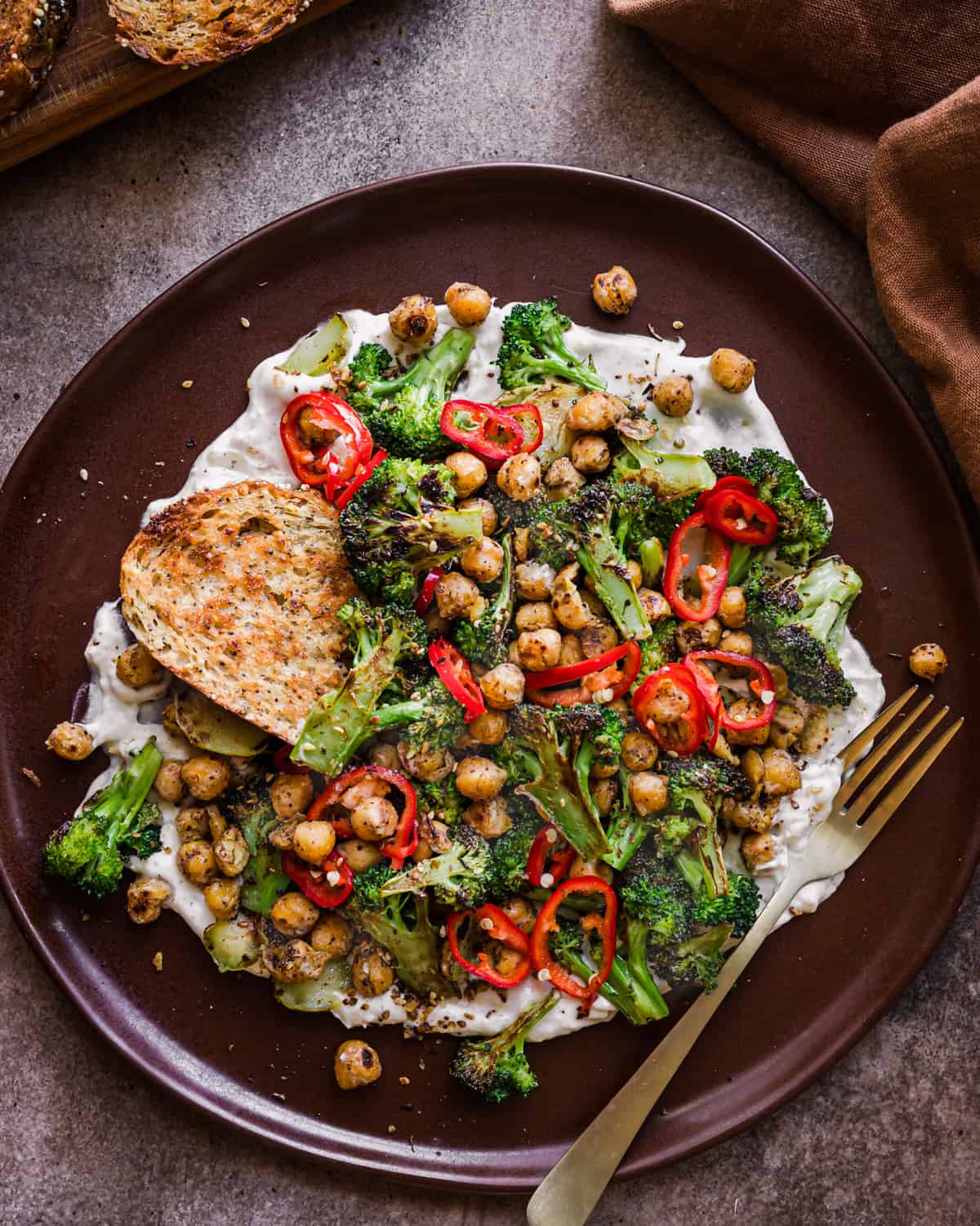 Charred broccoli and spiced chickpeas on yogurt sauce with bread on a brown plate with fork. 