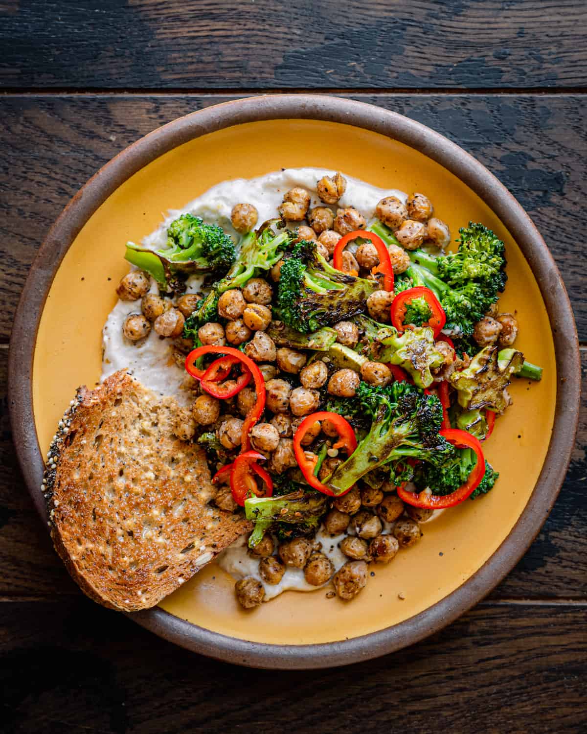 Spiced chickpeas and charred broccoli on yogurt sauce with bread on a yellow plate on a table.