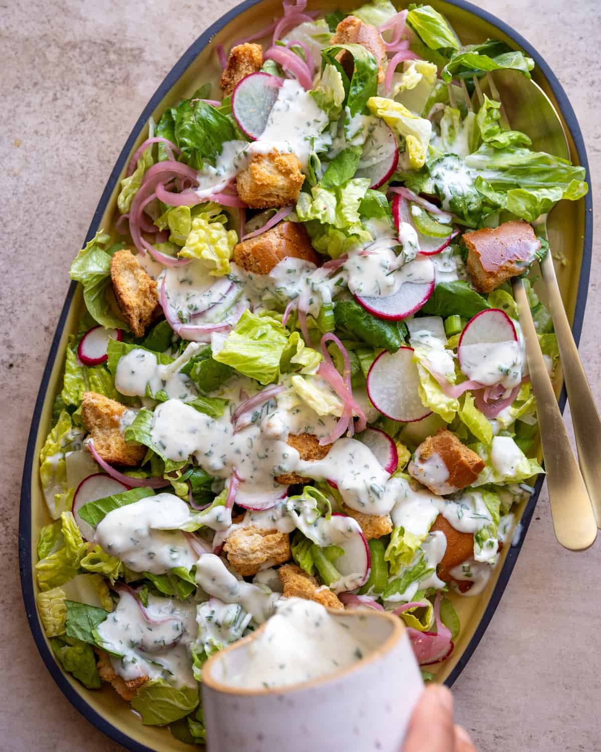 pouring vegan ranch dressing on top of romaine salad with croutons on a yellow platter. 