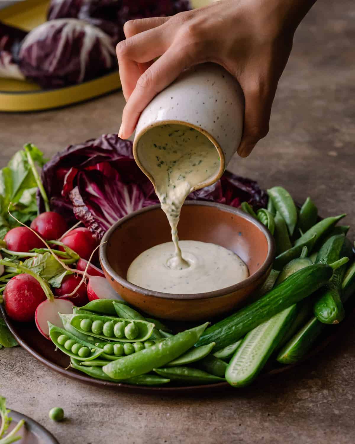 woman's hands pouring a vessel of vegan ranch dressing into a bowl in a crudites platter.