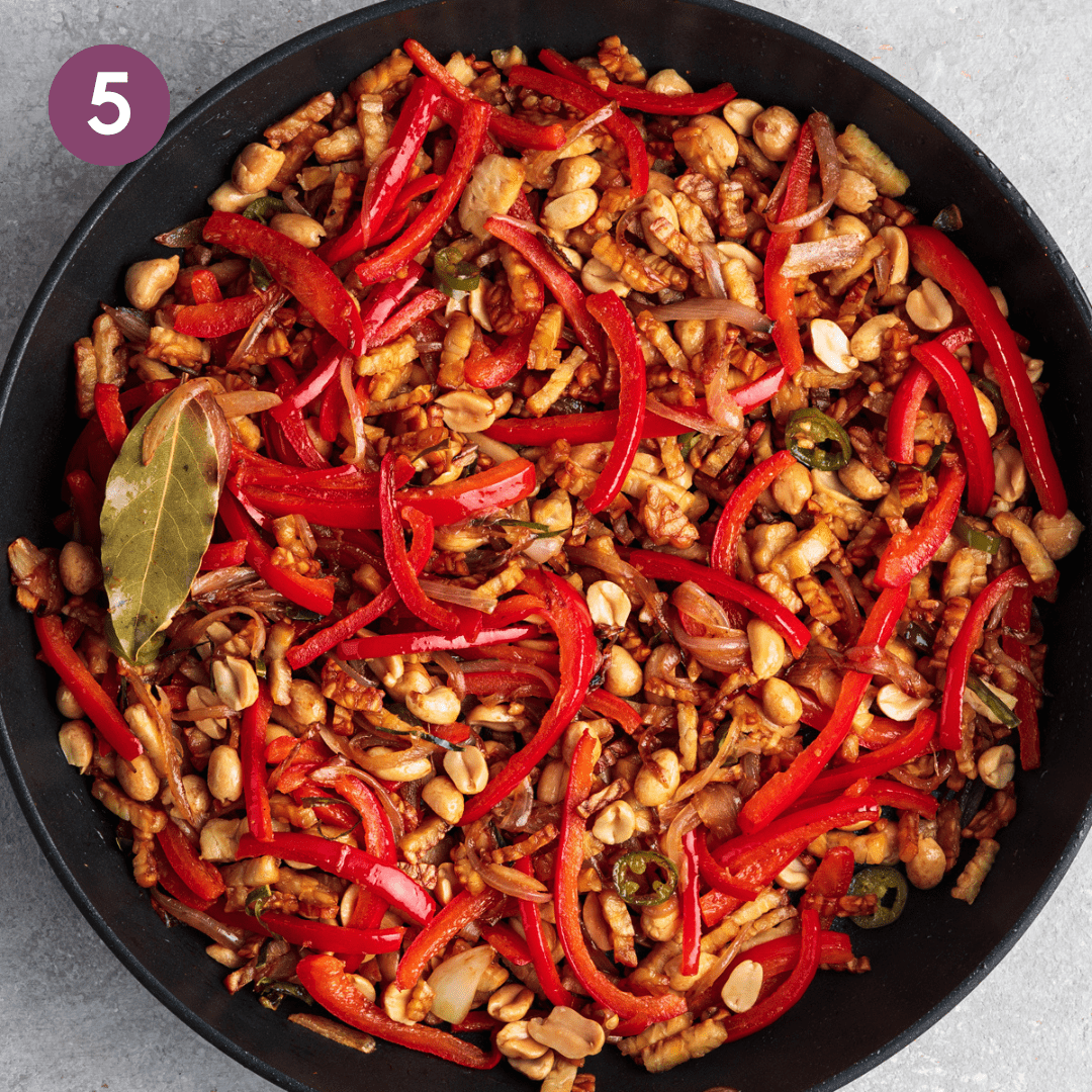 Tempeh and peanuts added to aromatics and bell peppers in frying pan.