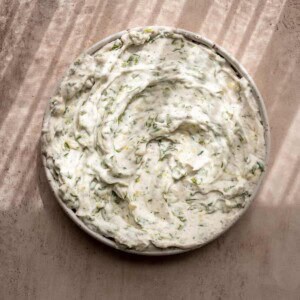 creamy vegan tzatziki swirled on a ceramic plate on a light pink surface with dappled light.