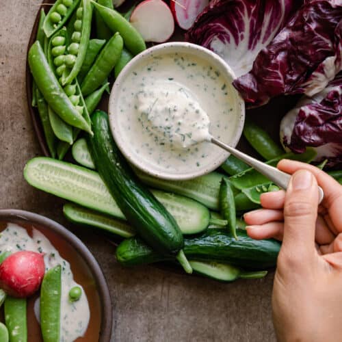 woman's hands dipping a spoon into bowl of vegan ranch dressing surrounded by crudites platter.