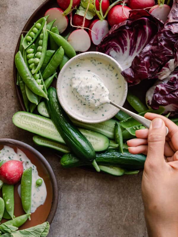 woman's hands dipping a spoon into bowl of vegan ranch dressing surrounded by crudites platter.