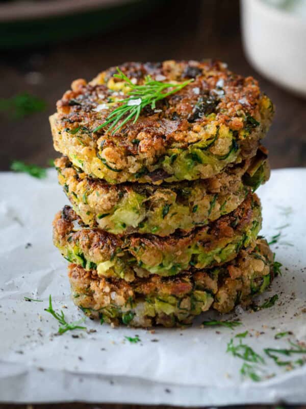 a stack of four vegan zucchini fritters on parchment paper with herbs and tzatziki on the side.