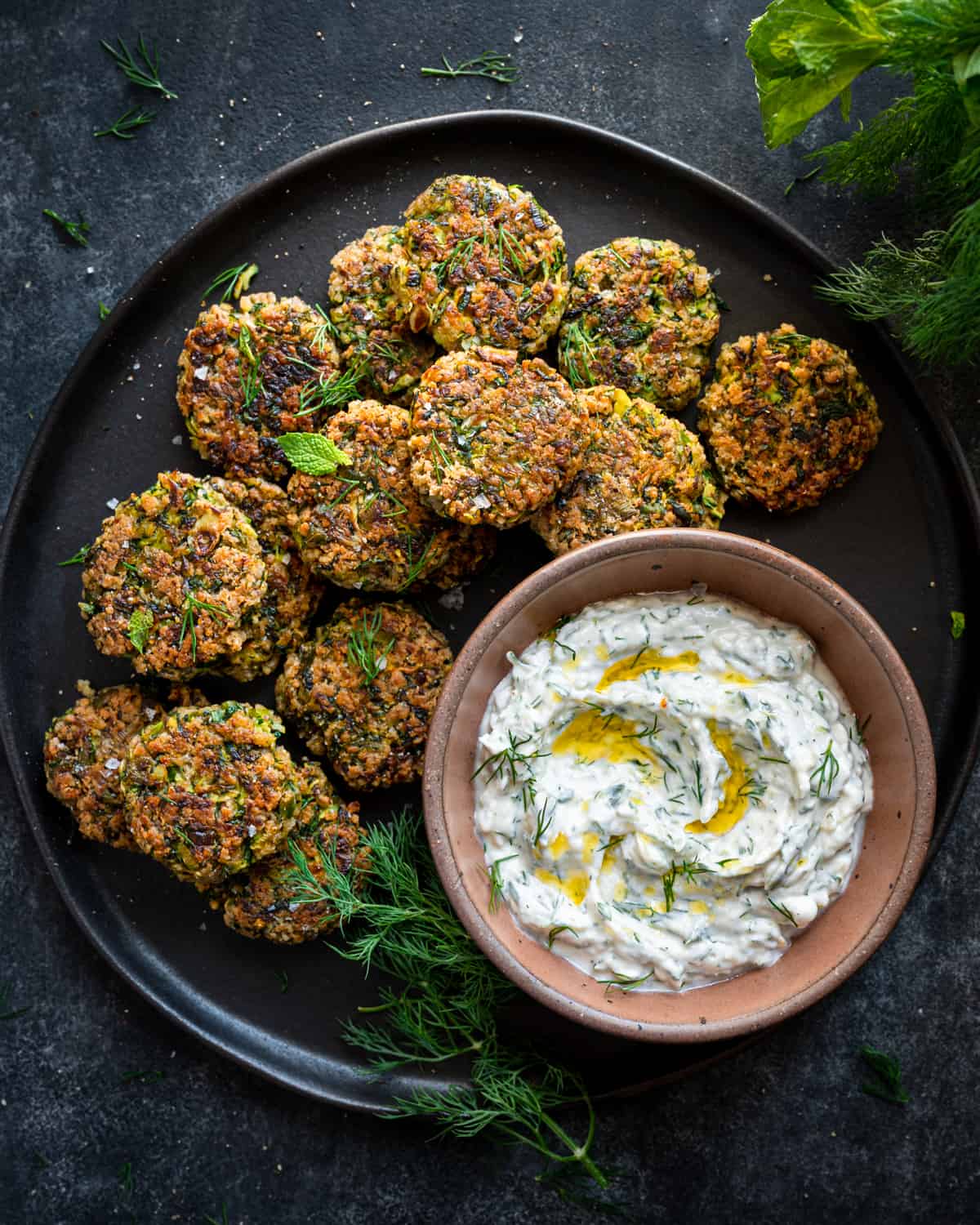 vegan zucchini fritters spread out on a black plate with a bowl of vegan tzatziki.