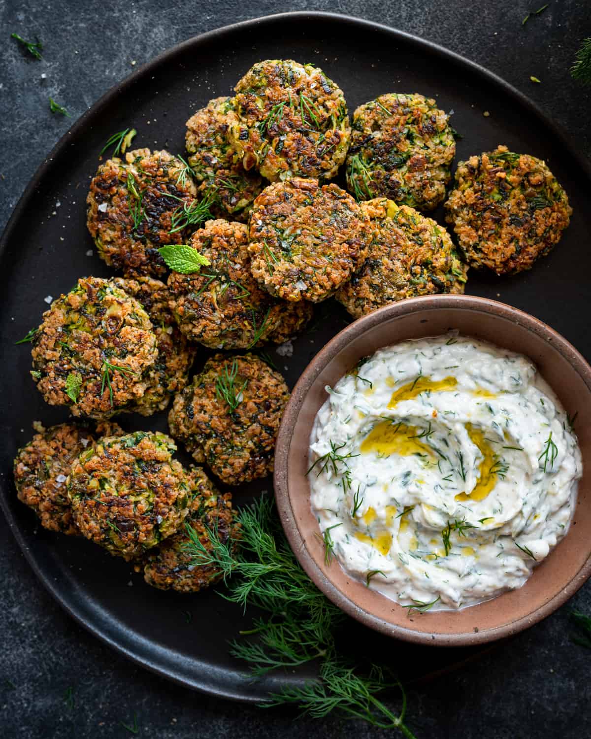 vegan zucchini fritters spread out on a black plate with a bowl of vegan tzatziki. 