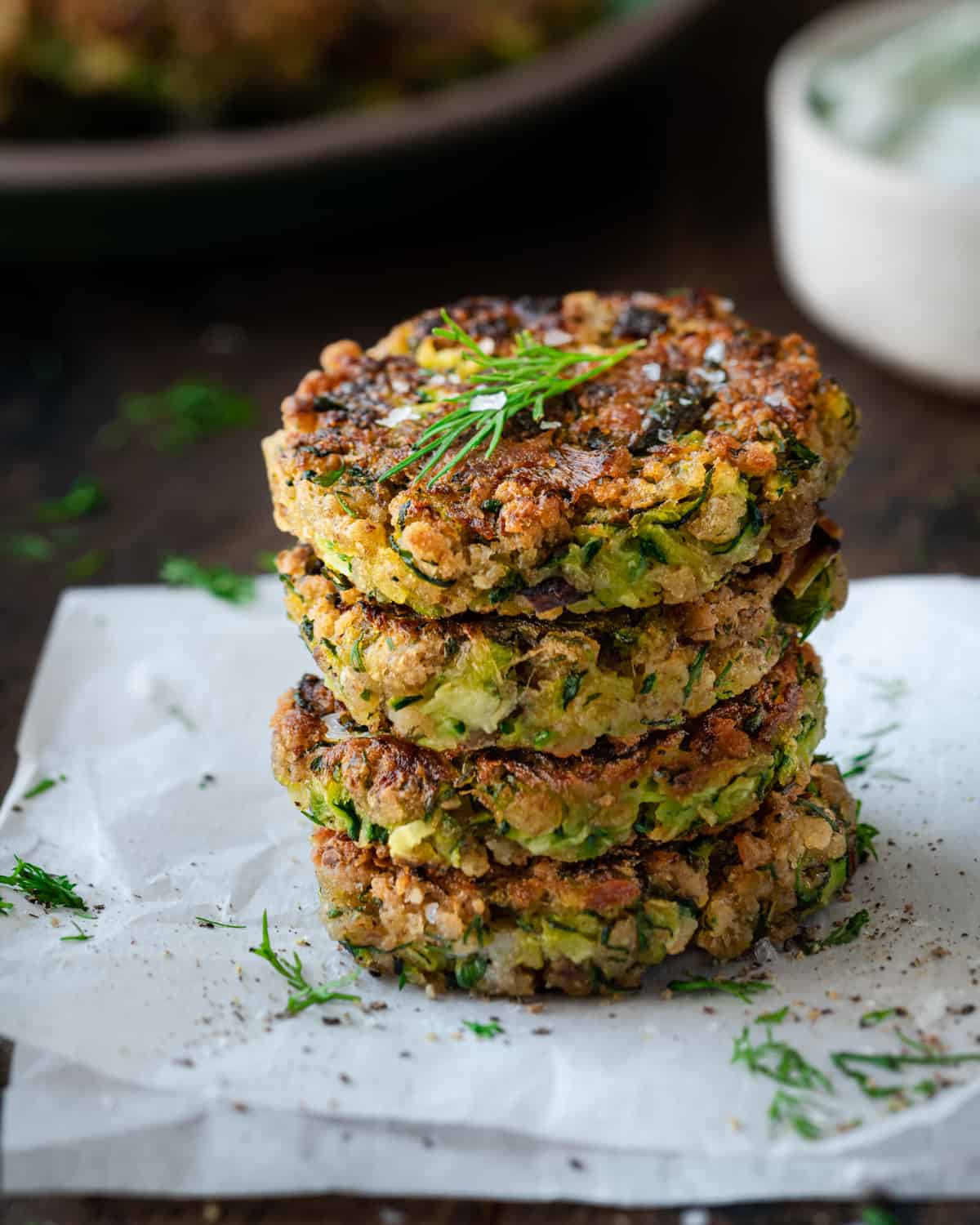 a stack of four vegan zucchini fritters on parchment paper with herbs and tzatziki on the side.