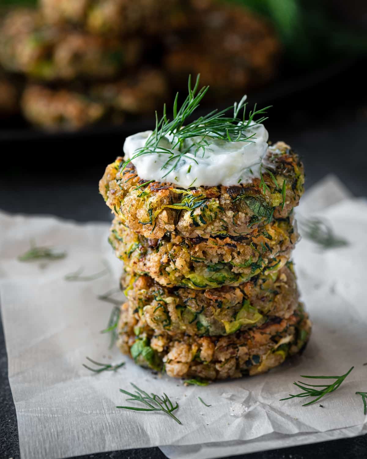 a stack of vegan zucchini fritters on parchment paper with vegan tzatziki and dill on top.