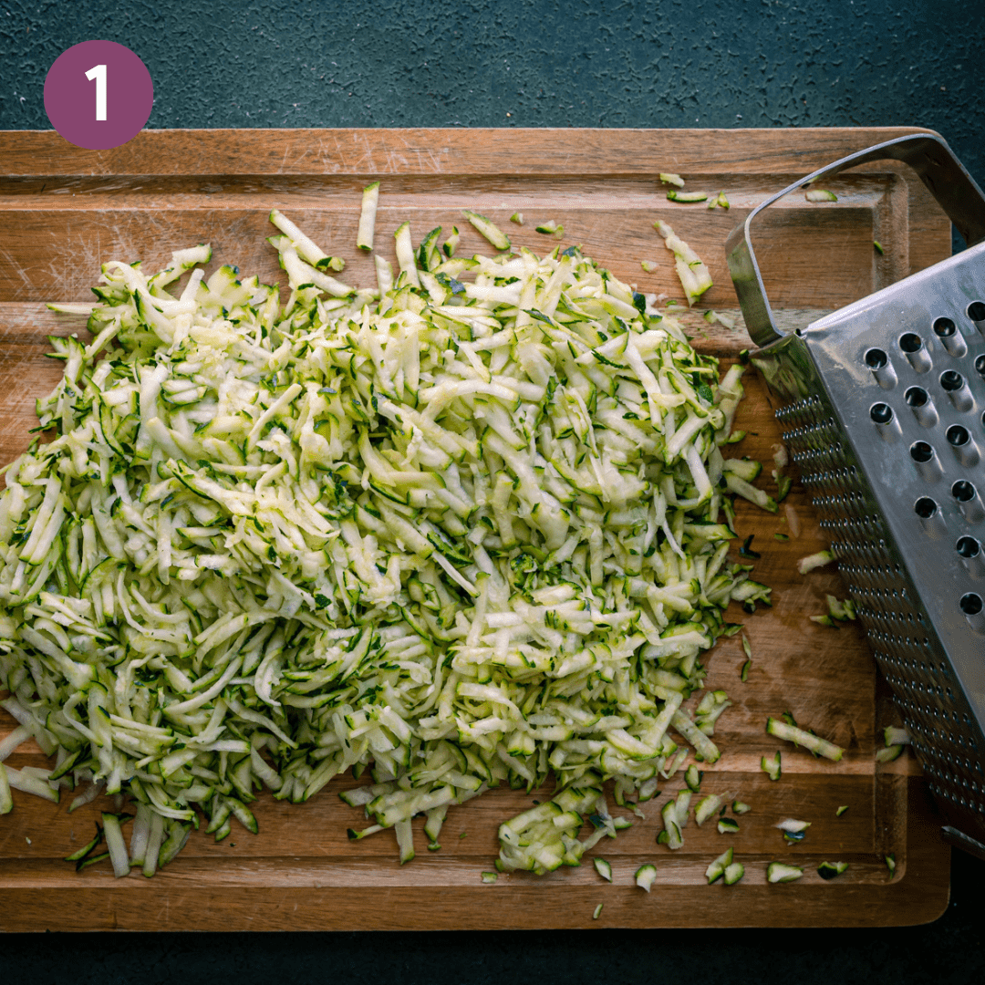 grated zucchini on a wooden cutting board next to a box grater. 