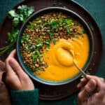 woman's hands digging a spoon into creamy butternut squash soup topped with lentils and herbs.