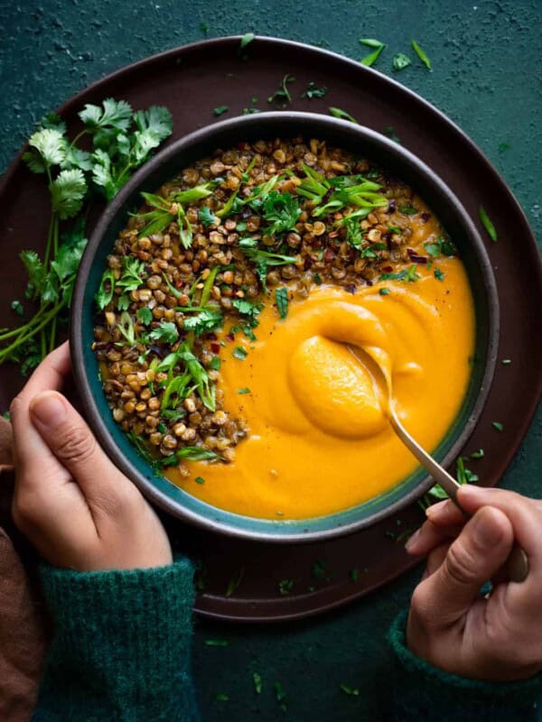 woman's hands digging a spoon into creamy butternut squash soup topped with lentils and herbs.
