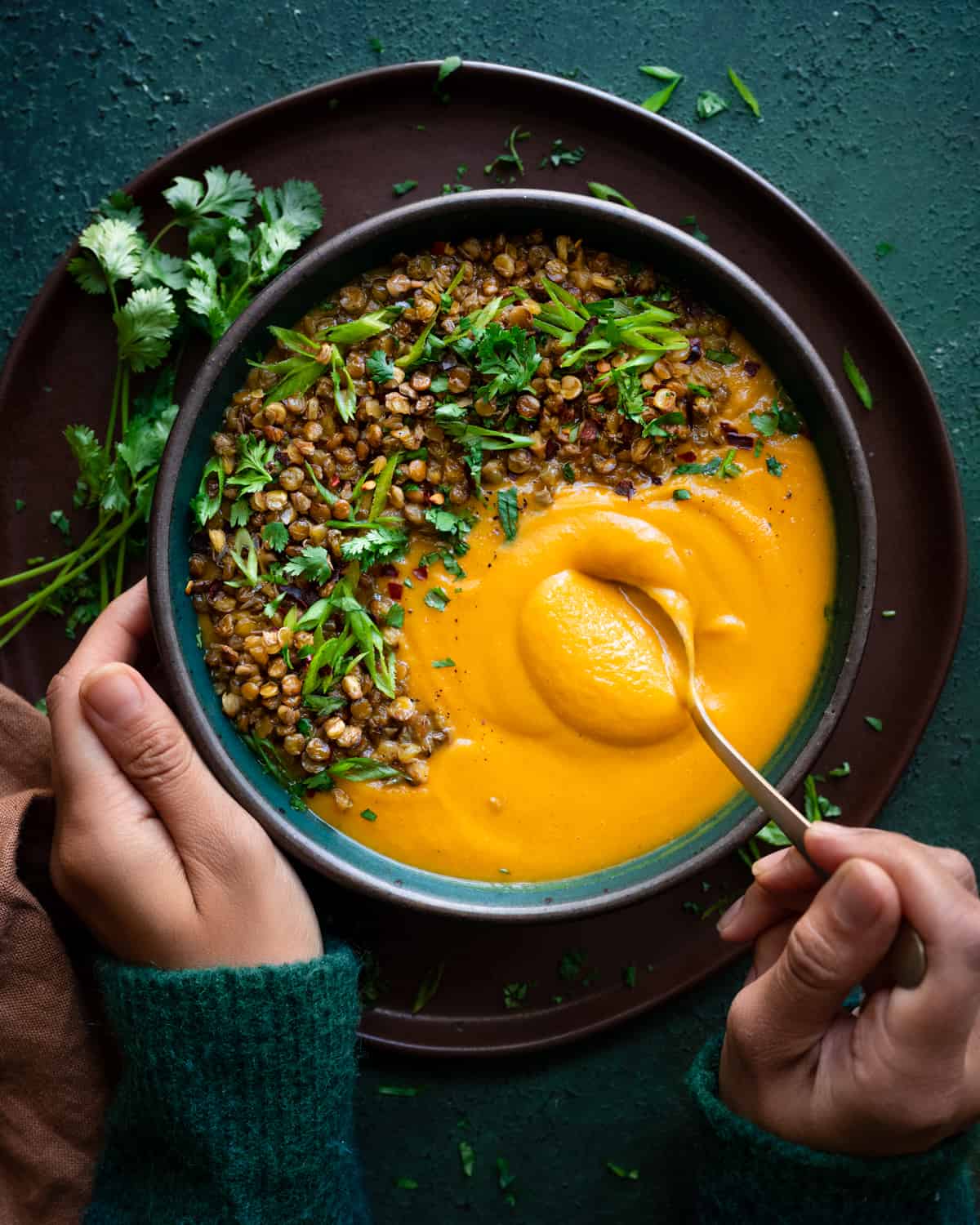 woman's hands digging a spoon into creamy butternut squash soup topped with lentils and herbs.