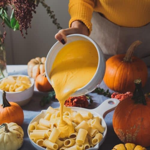 Woman pouring cheese sauce onto noodles in a large skillet on table next to various pumpkins.