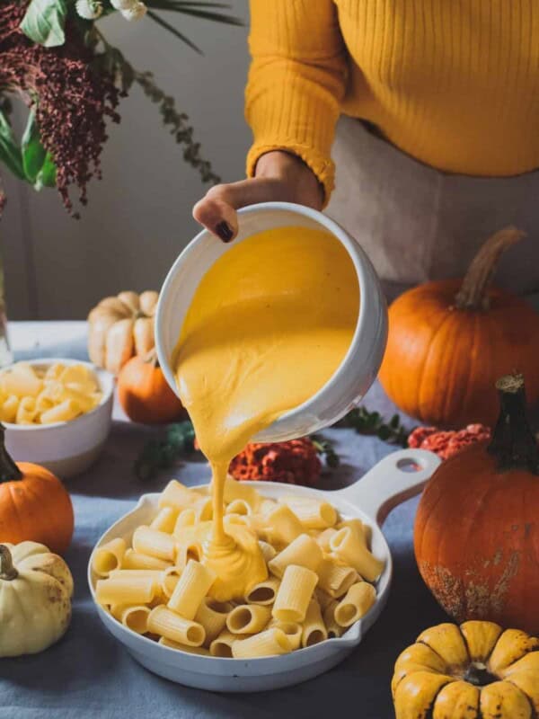 Woman pouring cheese sauce onto noodles in a large skillet on table next to various pumpkins.