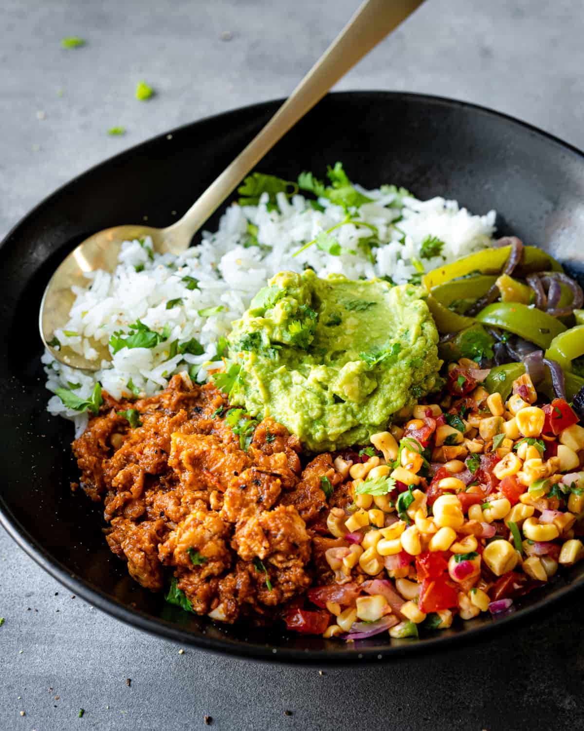 Vegan burrito bowl with sofritas, corn salsa, guacamole, and rice in a black ceramic bowl with spoon. 