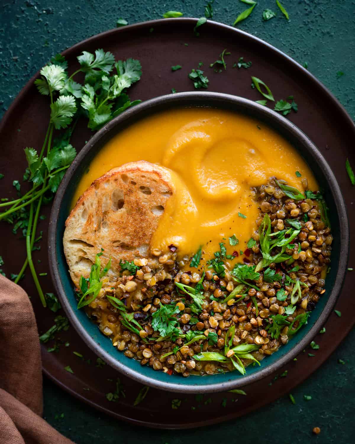 butternut squash soup topped with crispy lentils, fresh herbs, and served with bread in bowl on top of brown plate.