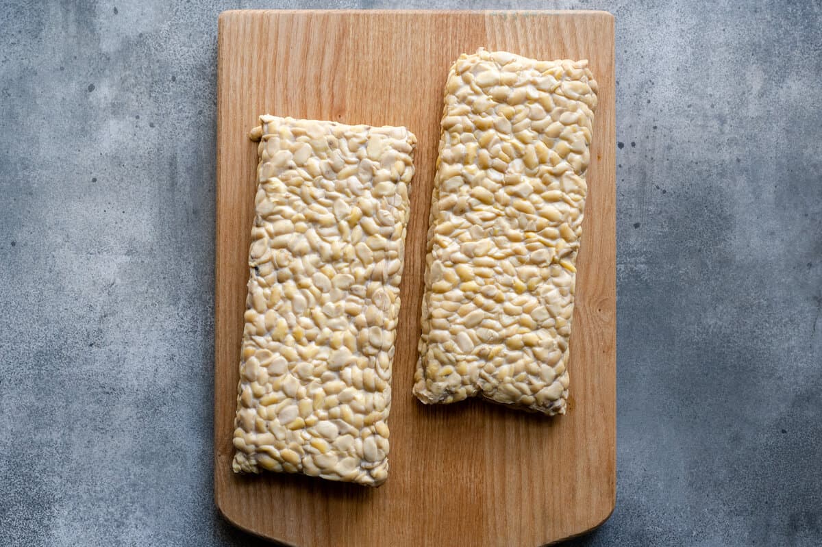 two blocks of tempeh on a wooden cutting board on a grey-blue surface.