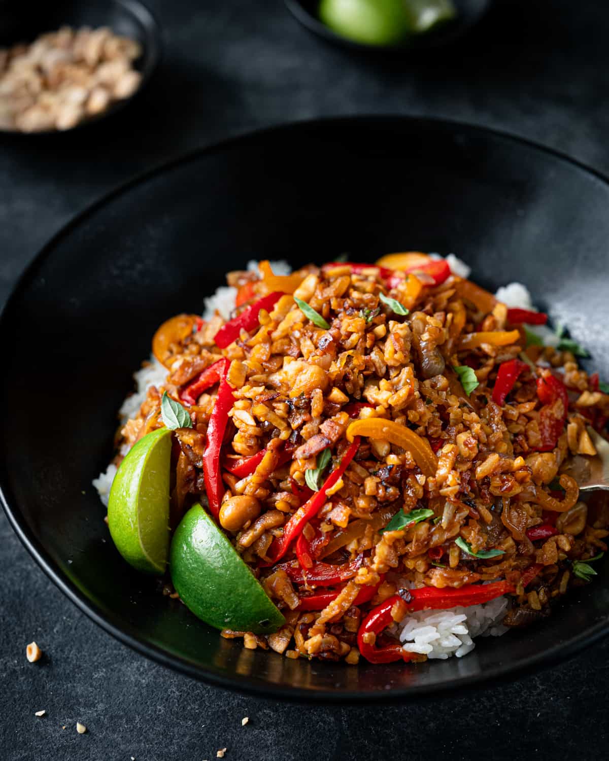 tempeh kecap stir fry in a black bowl over white rice with lime wedges.