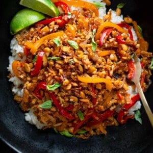 tempeh kecap stir fry with peanuts in a black bowl over white rice with lime wedges.