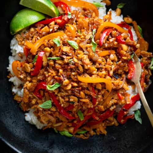 tempeh kecap stir fry with peanuts in a black bowl over white rice with lime wedges.