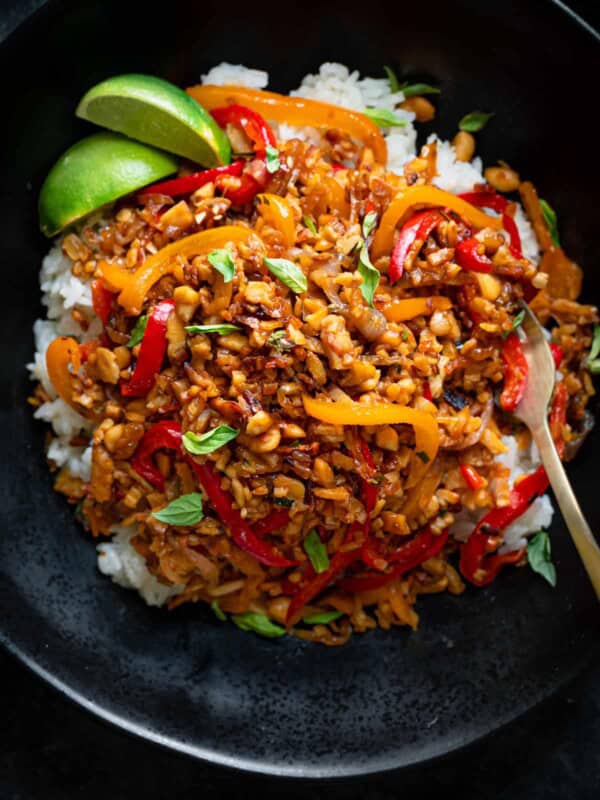 tempeh kecap stir fry with peanuts in a black bowl over white rice with lime wedges.